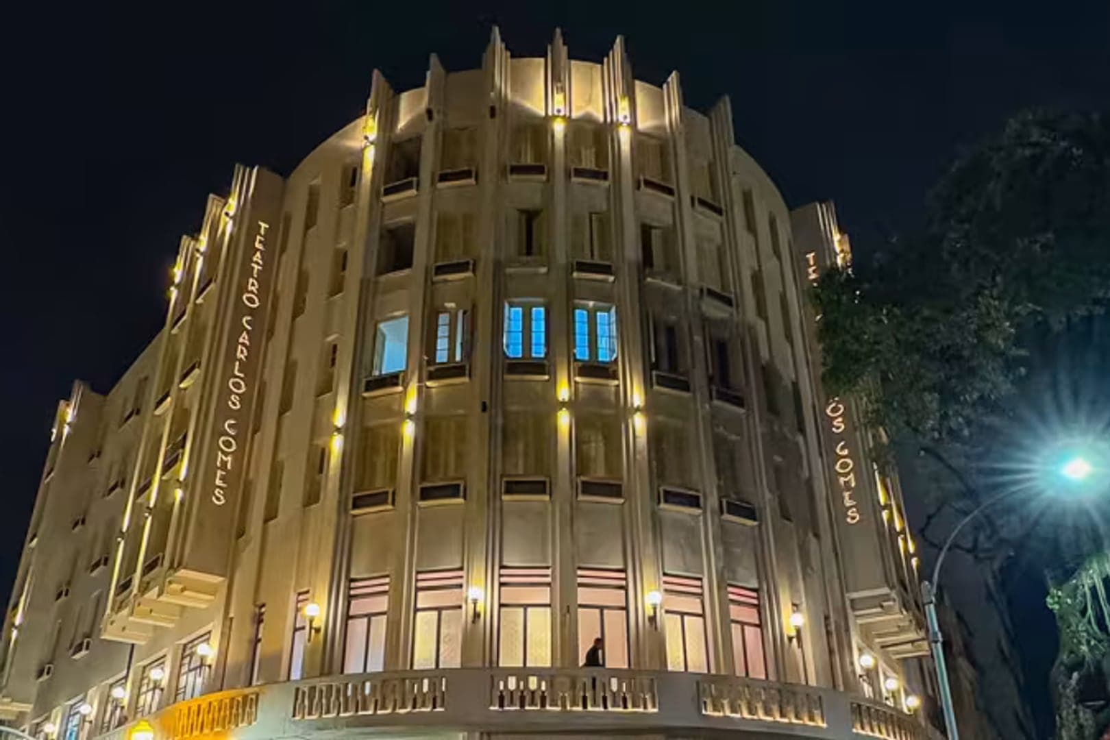 Fachada art déco do Teatro Municipal Carlos Gomes iluminada à noite, na Praça Tiradentes, com o nome do teatro em letras douradas na coluna